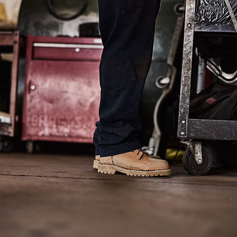 Person wearing tan work boots in a workshop setting with tools and equipment.