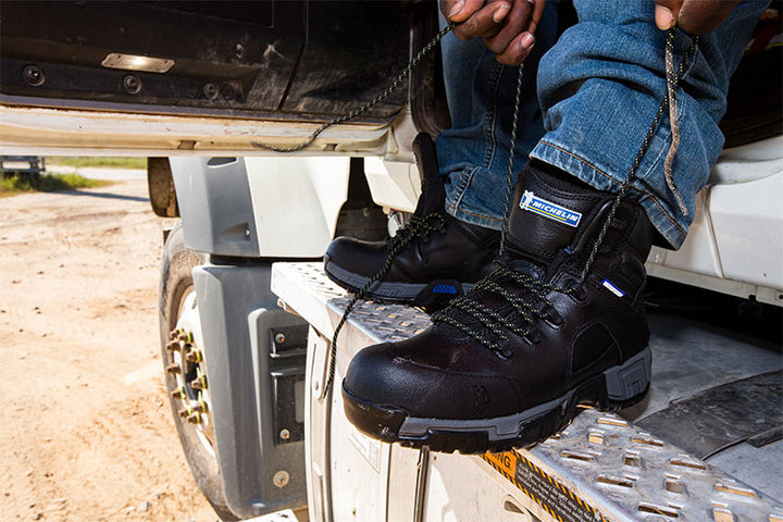Person wearing black work boots with visible branding, standing on a vehicle's bumper.